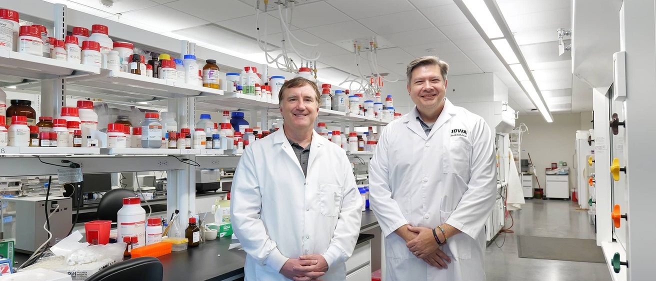 Two individuals in white lab coats stand in a bright research laboratory lined with shelves of chemical containers and scientific equipment.